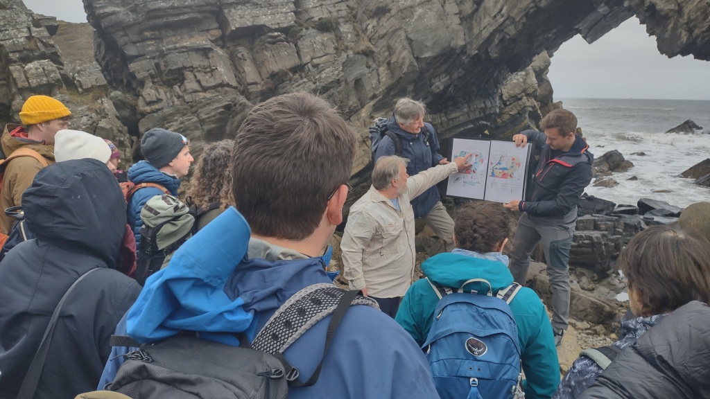 A group of geologists on a beach look towards someone explaining something on a map. The map is in a folder held by two other people. In the background is a rock arch. 